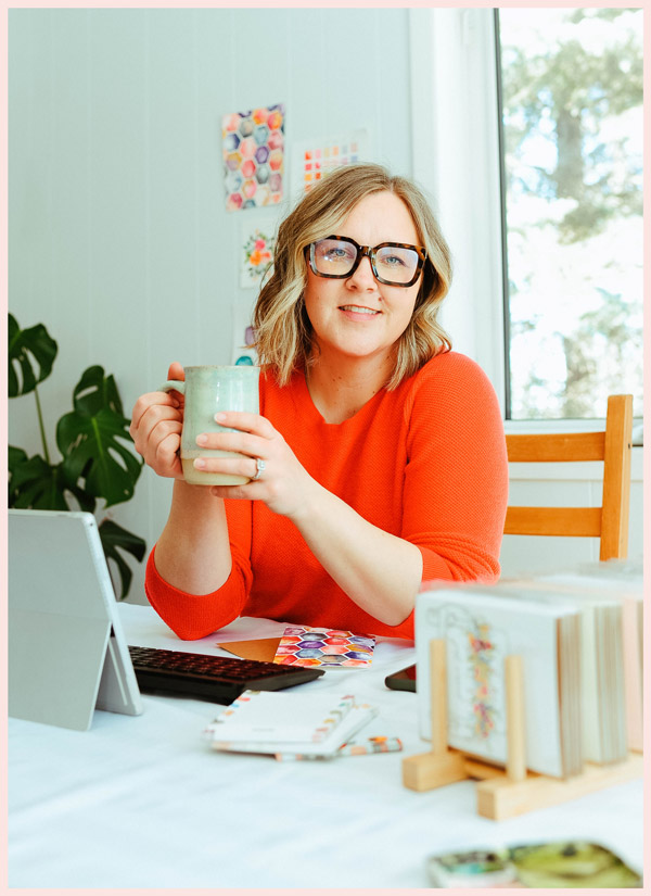 A woman in an orange shirt sits at a desk holding a coffee mug.