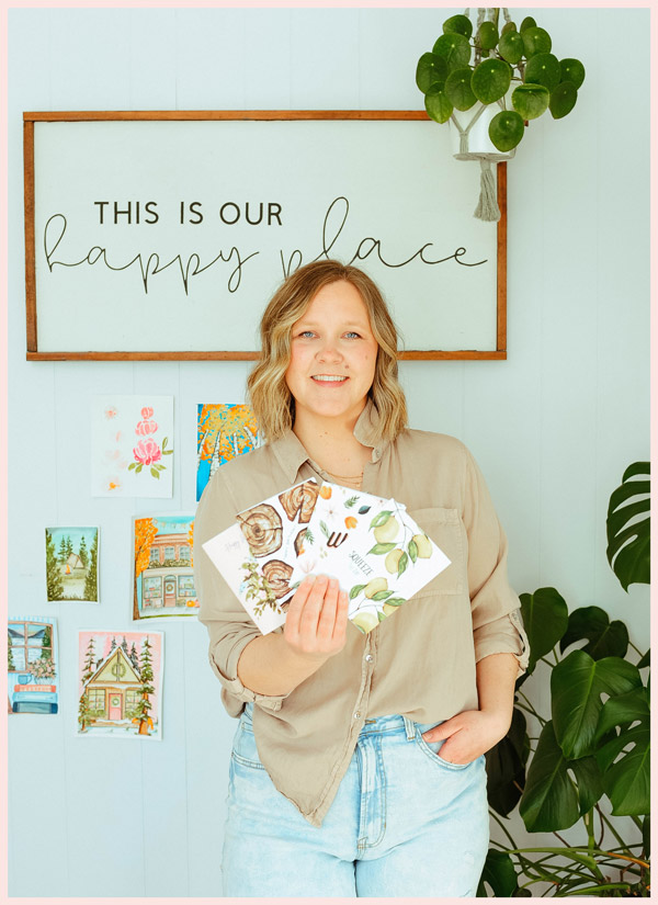 A woman holding some greeting cards stands in front of a sign that reads "this is our happy place".